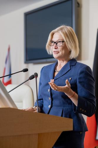 Content confident mature Caucasian business woman in suit standing at rostrum with microphones and gesturing hand while giving speech